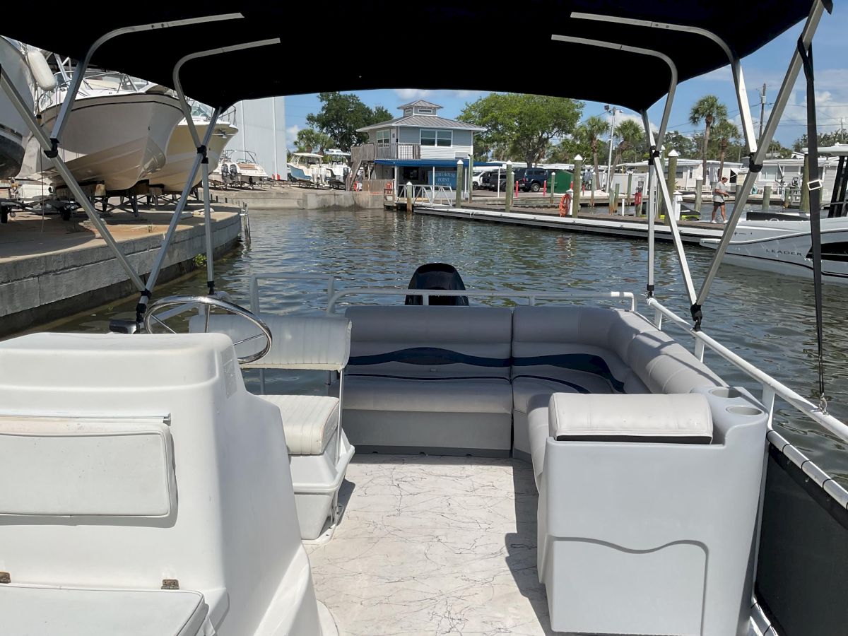 View from a pontoon boat showing cushioned seating, a calm marina canal, docks, and other boats with palm trees in the background today.