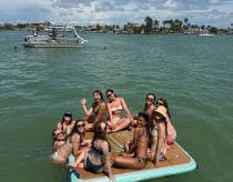 A group of swimmers ride a small inflatable raft in the ocean on a sunny day, with a coastline in the background.