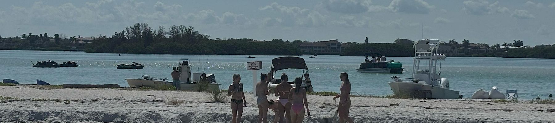 People walk on a sandy beach by the shoreline with several boats and jet skis anchored in calm blue water.