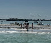 People walk on a sandy beach by the shoreline with several boats and jet skis anchored in calm blue water.