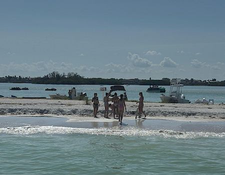 People walk on a sandy beach by the shoreline with several boats and jet skis anchored in calm blue water.