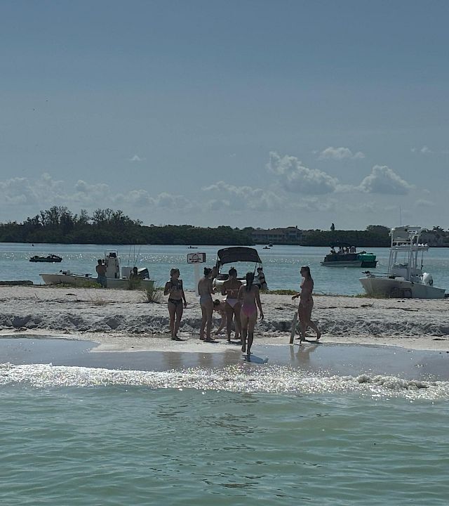 People walk on a sandy beach by the shoreline with several boats and jet skis anchored in calm blue water.