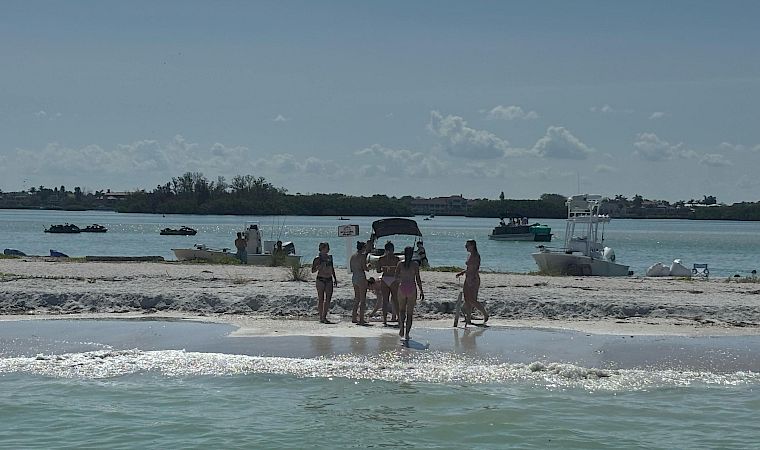 People walk on a sandy beach by the shoreline with several boats and jet skis anchored in calm blue water.