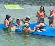 Friends on a blue raft float on a river, sipping drinks and sunbathing with a green inflatable board in the water.