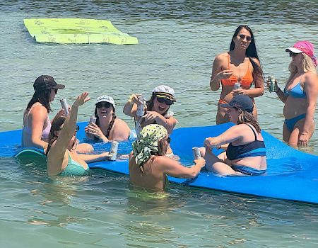 Friends on a blue raft float on a river, sipping drinks and sunbathing with a green inflatable board in the water.