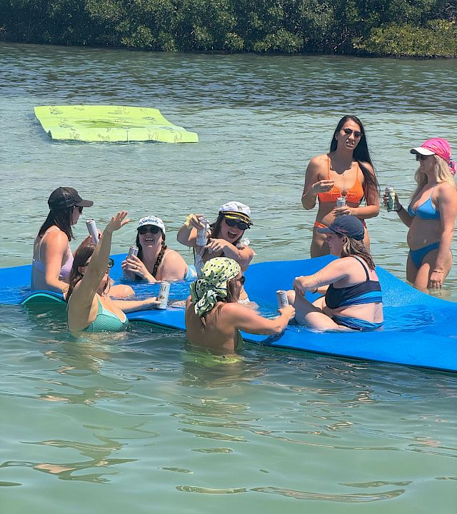 Friends on a blue raft float on a river, sipping drinks and sunbathing with a green inflatable board in the water.