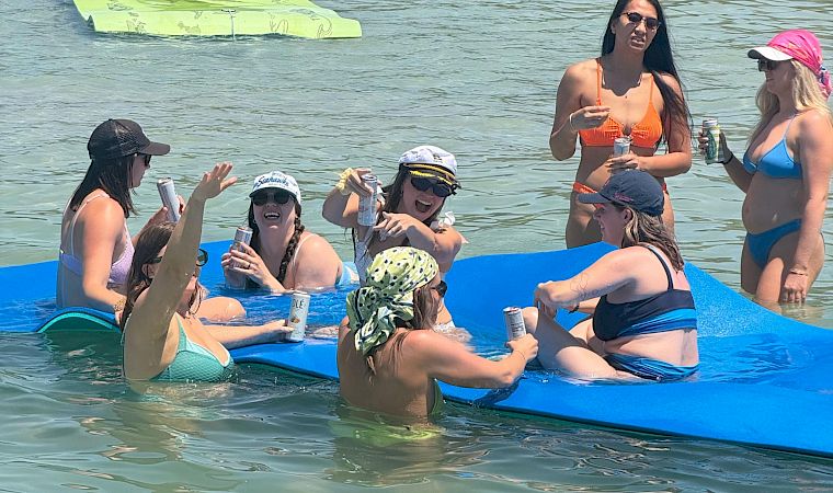 Friends on a blue raft float on a river, sipping drinks and sunbathing with a green inflatable board in the water.