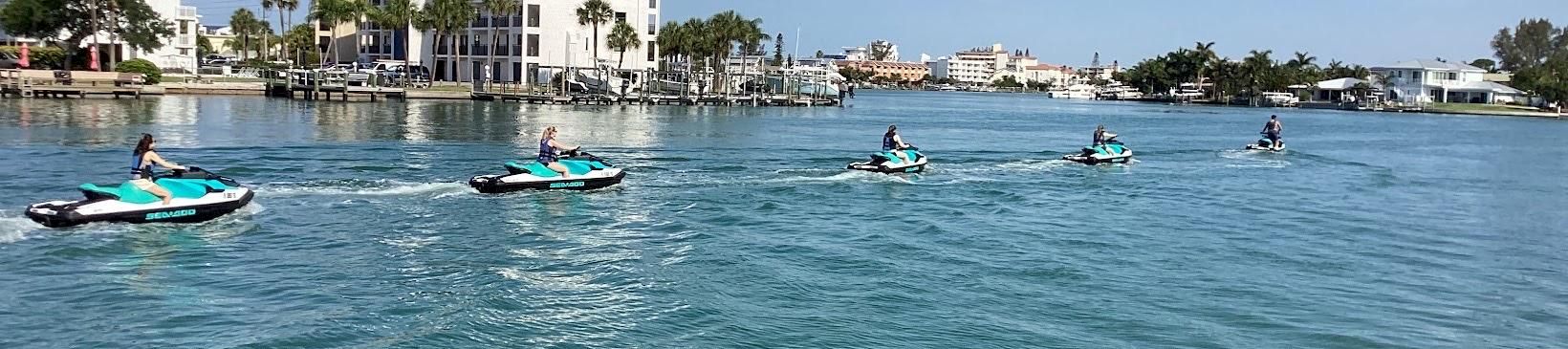 People ride jet skis across calm blue water beside a waterfront hotel and palm trees under a clear sky.