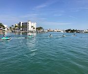 People ride jet skis across calm blue water beside a waterfront hotel and palm trees under a clear sky.