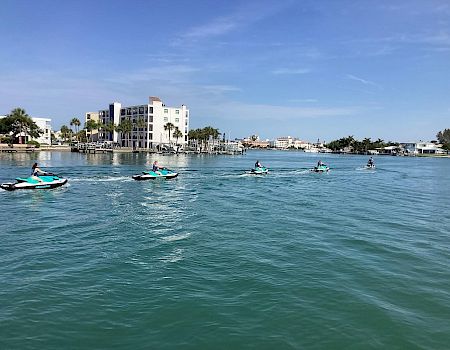 People ride jet skis across calm blue water beside a waterfront hotel and palm trees under a clear sky.
