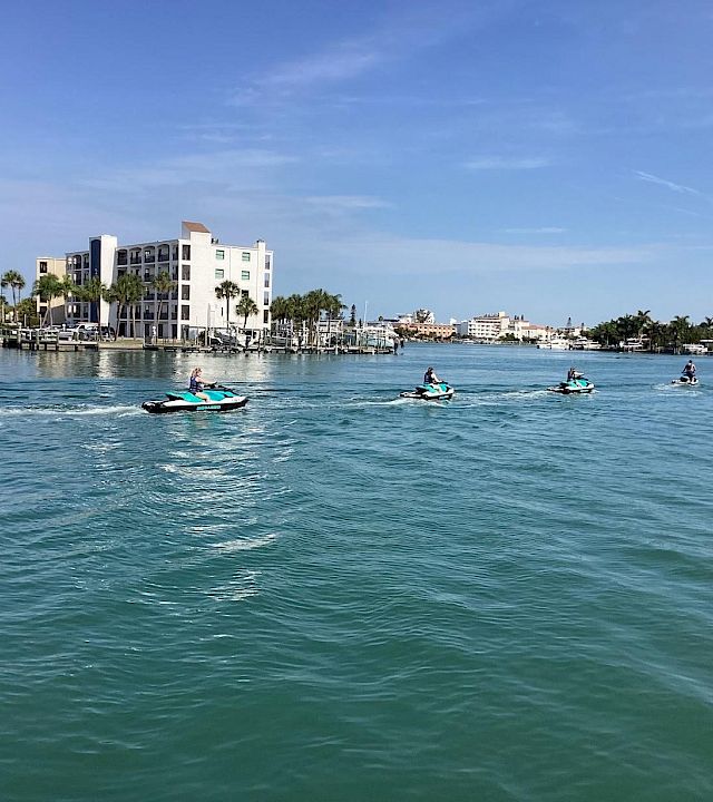 People ride jet skis across calm blue water beside a waterfront hotel and palm trees under a clear sky.