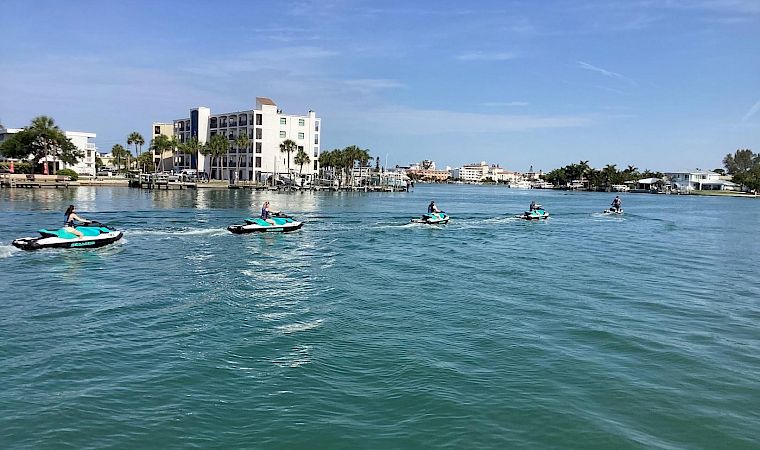 People ride jet skis across calm blue water beside a waterfront hotel and palm trees under a clear sky.