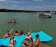 People in red swimsuits relax on a turquoise party pontoon boat in a sunny bay, with other anchored boats and clouds overhead.