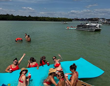 People in red swimsuits relax on a turquoise party pontoon boat in a sunny bay, with other anchored boats and clouds overhead.