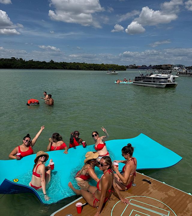People in red swimsuits relax on a turquoise party pontoon boat in a sunny bay, with other anchored boats and clouds overhead.