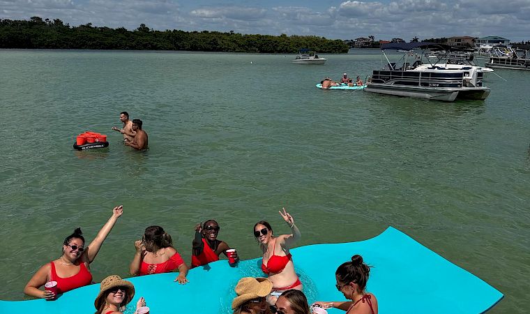 People in red swimsuits relax on a turquoise party pontoon boat in a sunny bay, with other anchored boats and clouds overhead.