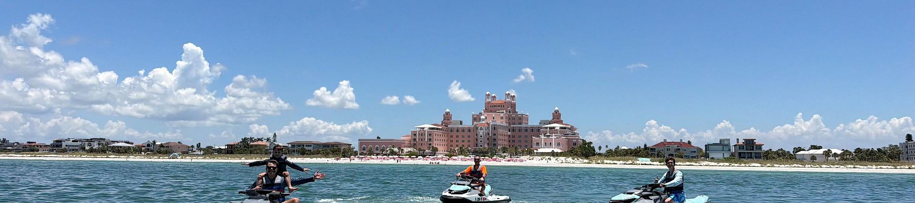 People ride jet skis on a sunny sea in front of a luxury hotel by the beach, with a blue sky and clouds.