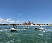 People ride jet skis on a sunny sea in front of a luxury hotel by the beach, with a blue sky and clouds.