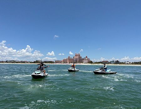 People ride jet skis on a sunny sea in front of a luxury hotel by the beach, with a blue sky and clouds.