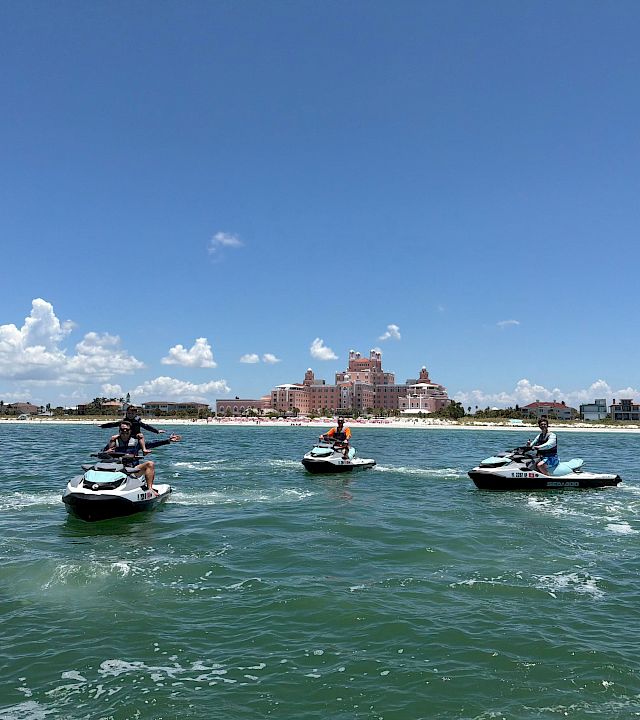 People ride jet skis on a sunny sea in front of a luxury hotel by the beach, with a blue sky and clouds.