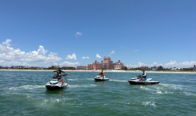 People ride jet skis on a sunny sea in front of a luxury hotel by the beach, with a blue sky and clouds.