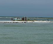 A small boat and people sit in the shallow surf on a windy beach under a gray sky.