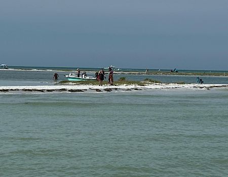 A small boat and people sit in the shallow surf on a windy beach under a gray sky.