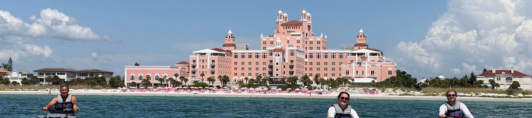 Jet skis speed across the water in front of a seaside pink hotel as three people take in the sunny sky.