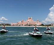 Jet skis speed across the water in front of a seaside pink hotel as three people take in the sunny sky.