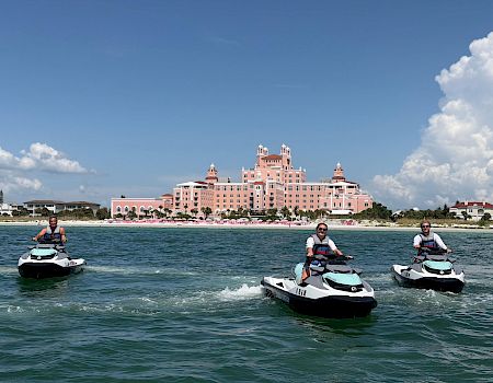 Jet skis speed across the water in front of a seaside pink hotel as three people take in the sunny sky.
