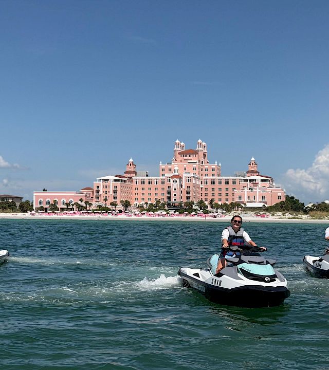 Jet skis speed across the water in front of a seaside pink hotel as three people take in the sunny sky.