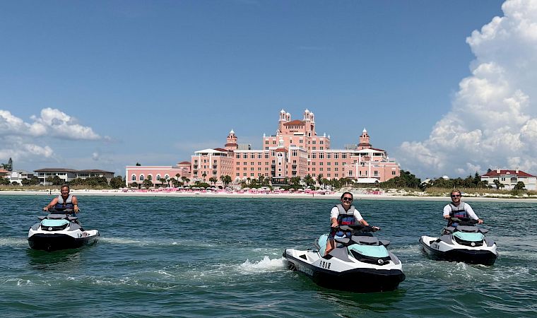 Jet skis speed across the water in front of a seaside pink hotel as three people take in the sunny sky.