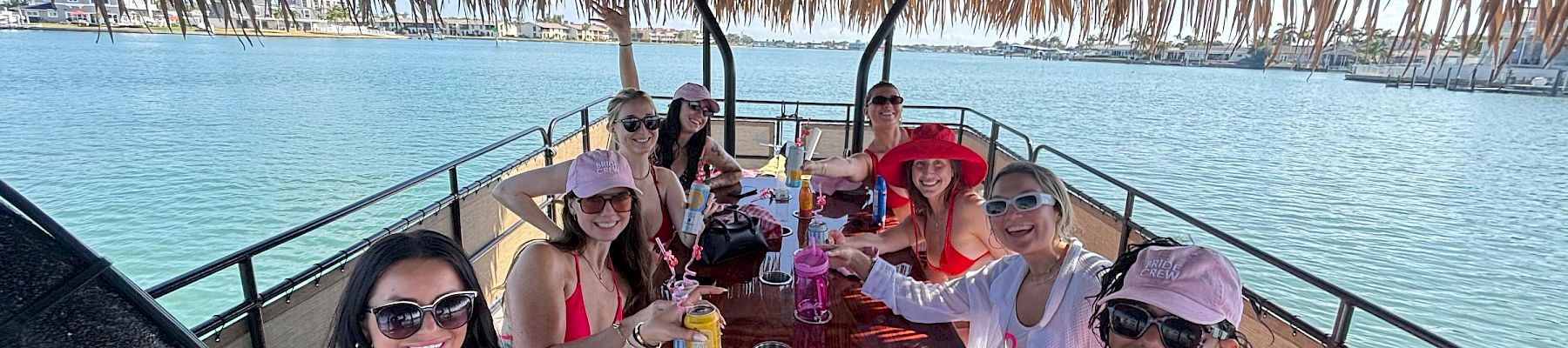 A group of friends on a boat party, cheering and toasting drinks under a tiki-style canopy by the sunny waterfront.