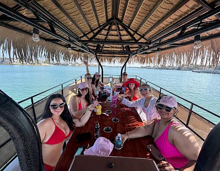 A group of friends on a boat party, cheering and toasting drinks under a tiki-style canopy by the sunny waterfront.