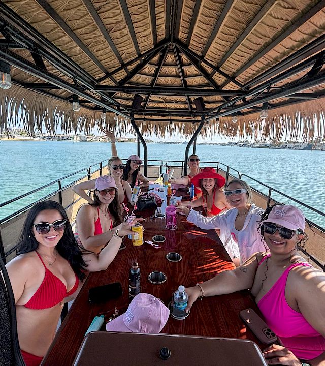 A group of friends on a boat party, cheering and toasting drinks under a tiki-style canopy by the sunny waterfront.