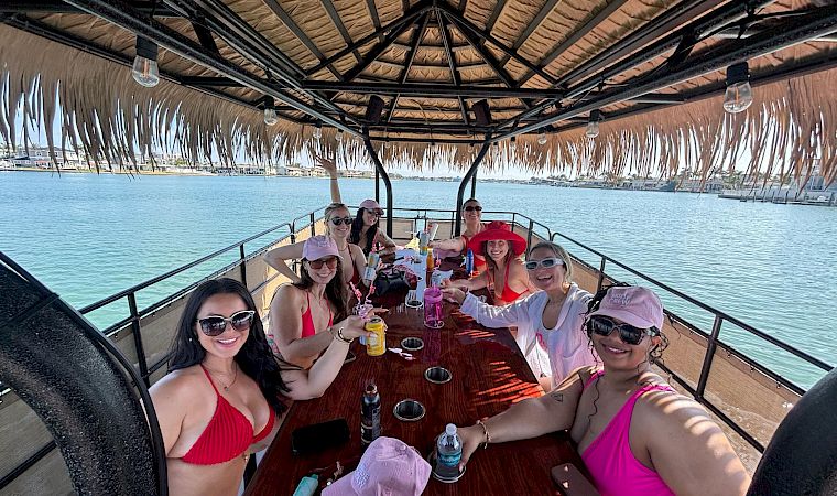 A group of friends on a boat party, cheering and toasting drinks under a tiki-style canopy by the sunny waterfront.
