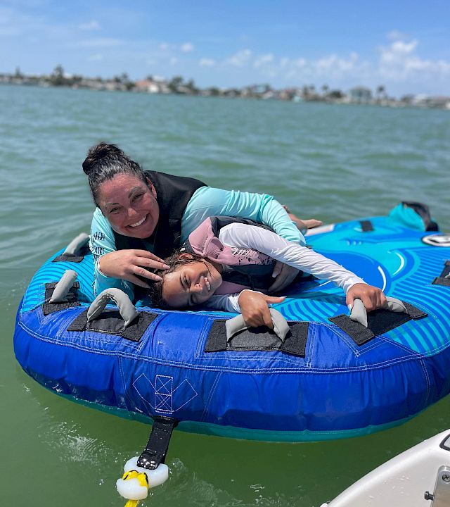 Two friends smile while riding a blue inflatable tube on the ocean in the sunshine, with a distant shoreline behind them.
