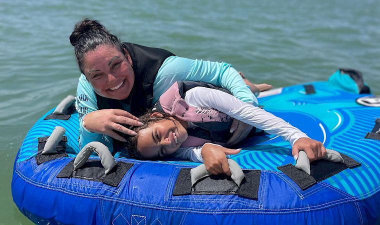Two friends smile while riding a blue inflatable tube on the ocean in the sunshine, with a distant shoreline behind them.