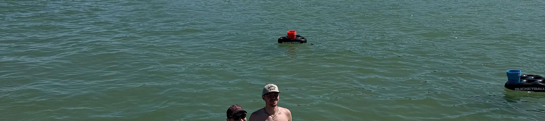 Friends relax on a sunny paddleboard/raft on a green river, cheering with drinks while heading toward a tree-lined shore.