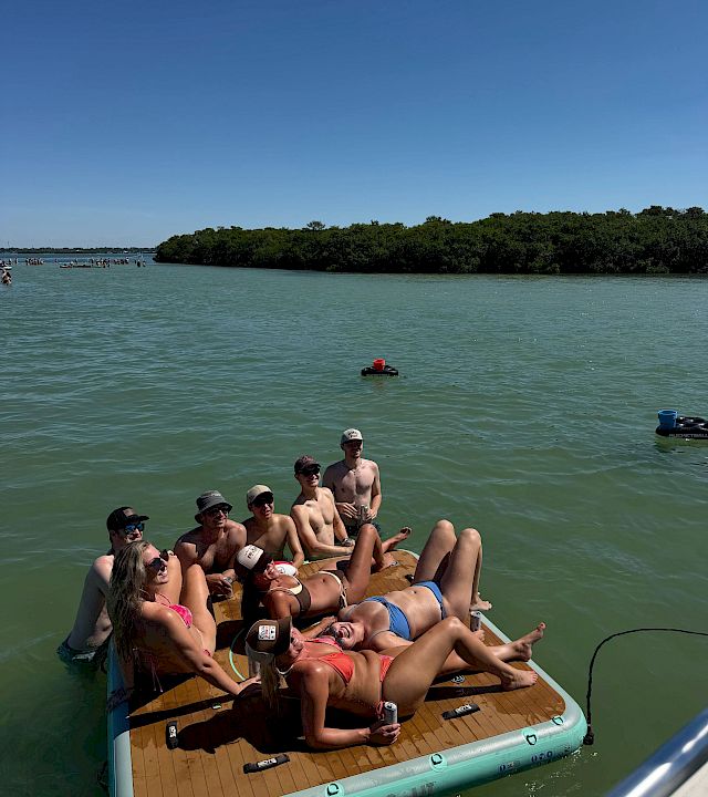 Friends relax on a sunny paddleboard/raft on a green river, cheering with drinks while heading toward a tree-lined shore.