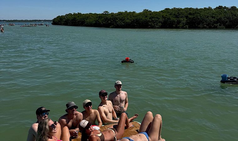 Friends relax on a sunny paddleboard/raft on a green river, cheering with drinks while heading toward a tree-lined shore.