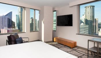 A modern hotel room with a large bed, wall-mounted TV, armchair, and cityscape views through large windows.