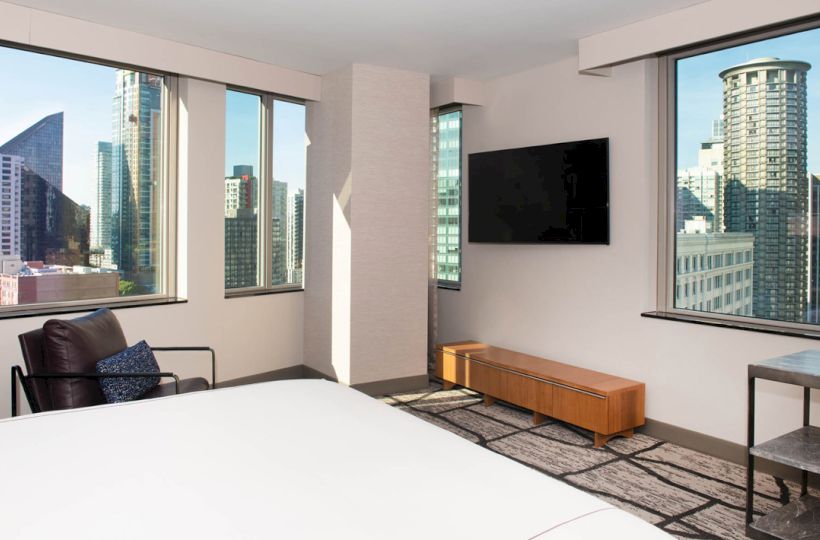 A modern hotel room with a large bed, wall-mounted TV, armchair, and cityscape views through large windows.