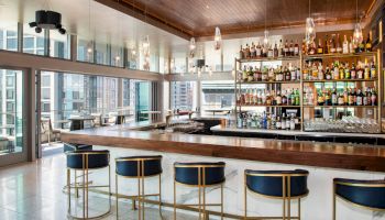 A modern bar with a sleek countertop, high stools, and a well-stocked liquor shelf, surrounded by large windows offering city views.
