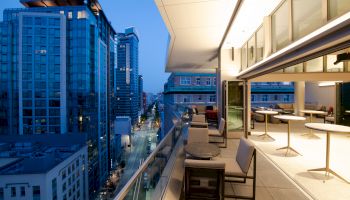A modern rooftop terrace overlooking a city street, with tables, chairs, and tall buildings visible during the evening.