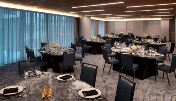 The image shows an elegantly set banquet hall with round tables, chairs, floral centerpieces, and soft lighting, prepared for an event.