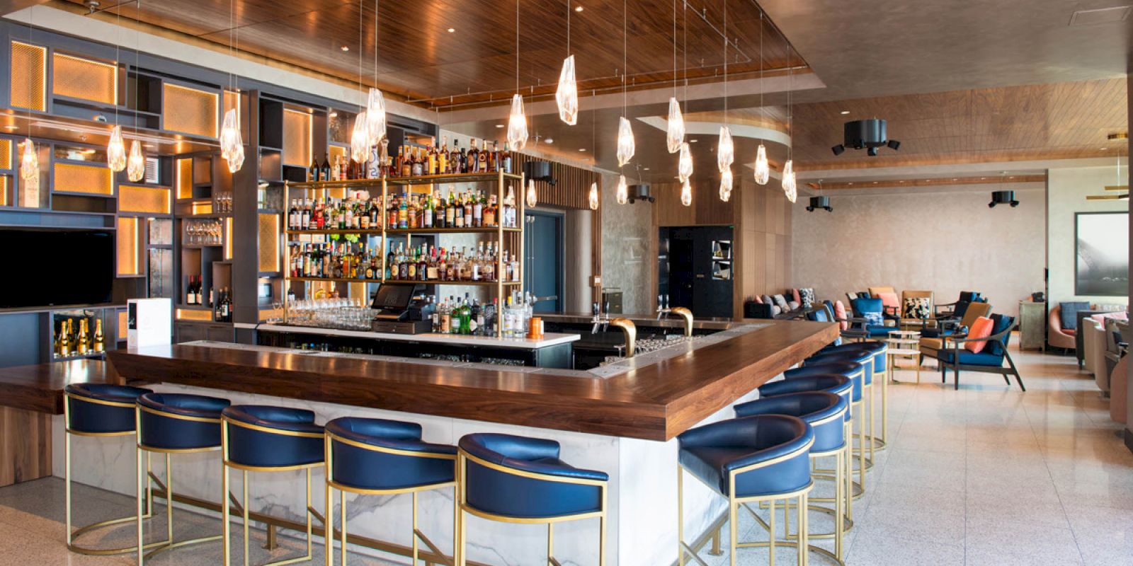 This image shows a modern bar with blue stools, hanging lights, and a well-stocked shelf. There are lounge areas and a large bar counter.