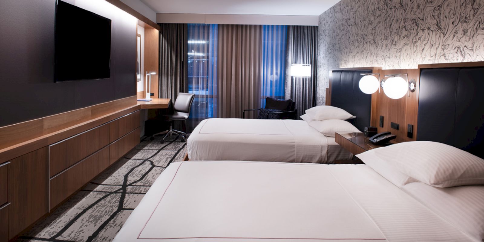 The image shows a modern hotel room with two neatly made beds, a TV, a desk, and a window with curtains partially open.