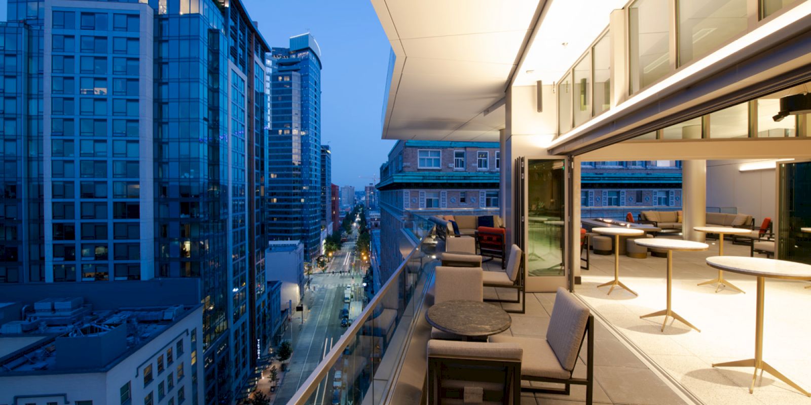 A modern balcony overlooking a cityscape at twilight, featuring sleek furniture and large glass windows with soft interior lighting.
