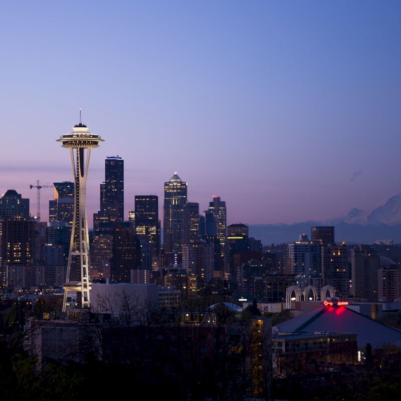 A city skyline at dusk with a prominent tower and distant mountain, featuring city lights and a clear sky transitioning to night.